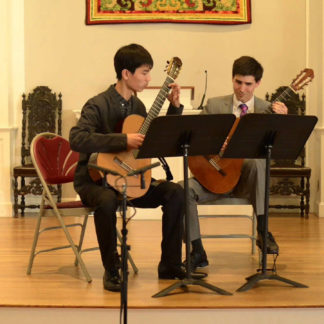 Image of a young man at left and a man at right, both holding guitars behind music stands.